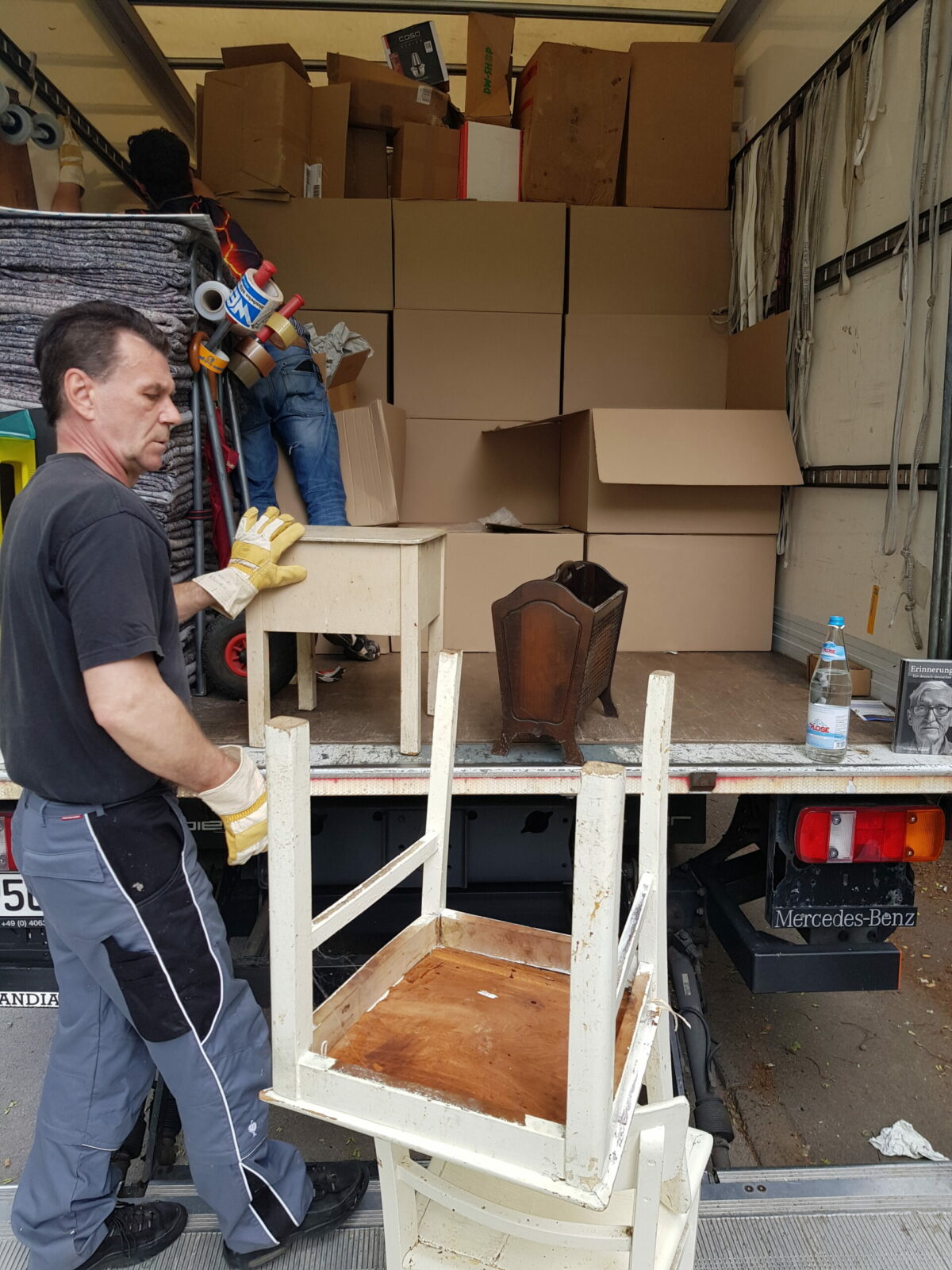 Mann lädt Möbel und Kartons in Umzugswagen, ein umgedrehter Stuhl und Tisch stehen auf der Ladebühne.