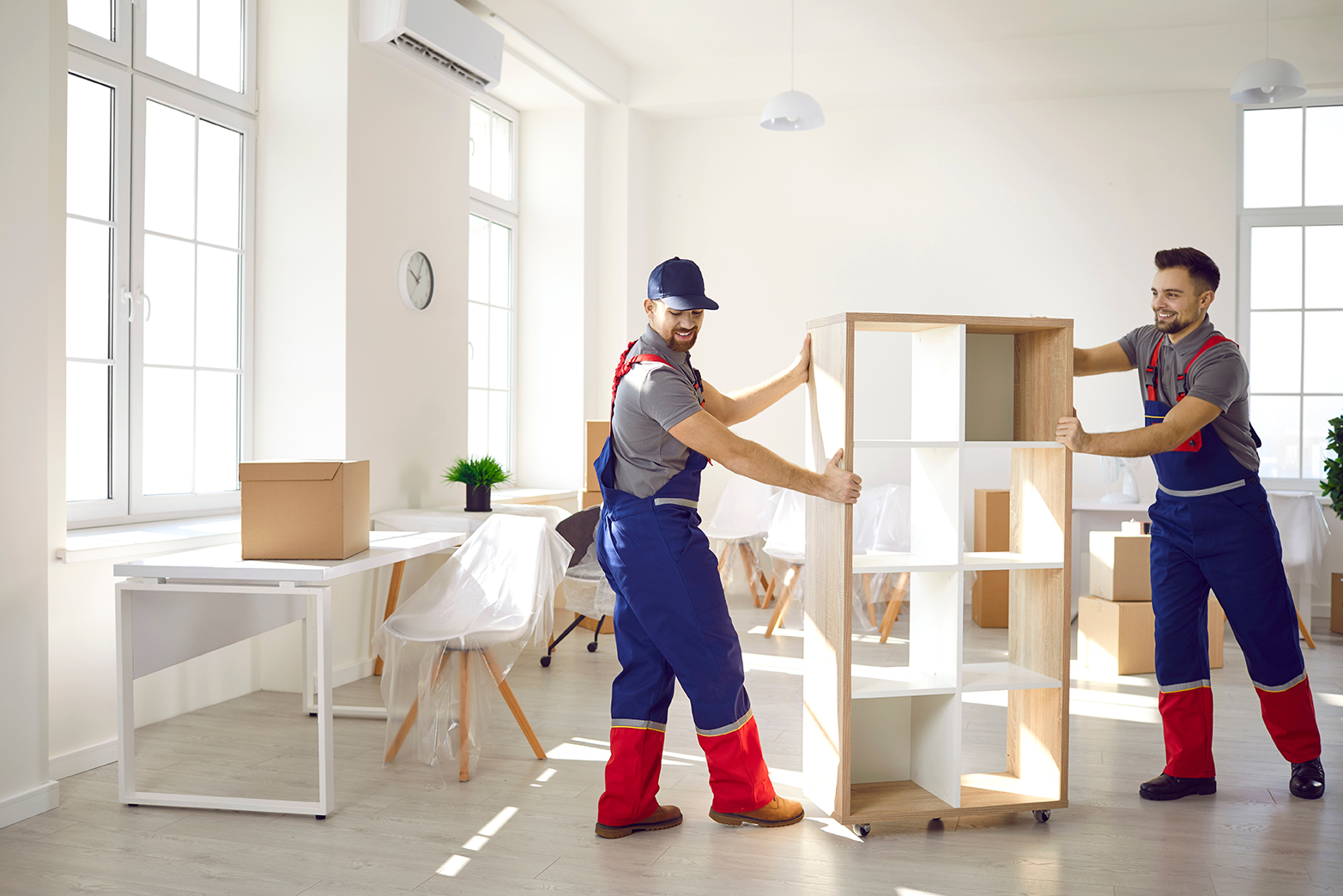 Zwei Umzugshelfer tragen ein Holzregal durch einen hellen, leeren Raum mit Kartons und abgedeckten Möbeln.