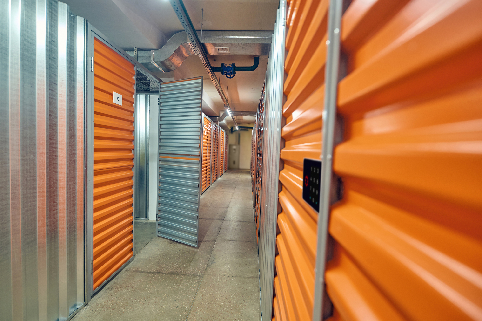 Flur in einem Lager mit orangefarbenen Metalltüren, eine Tür ist teilweise geöffnet, Rohre verlaufen an der Decke.